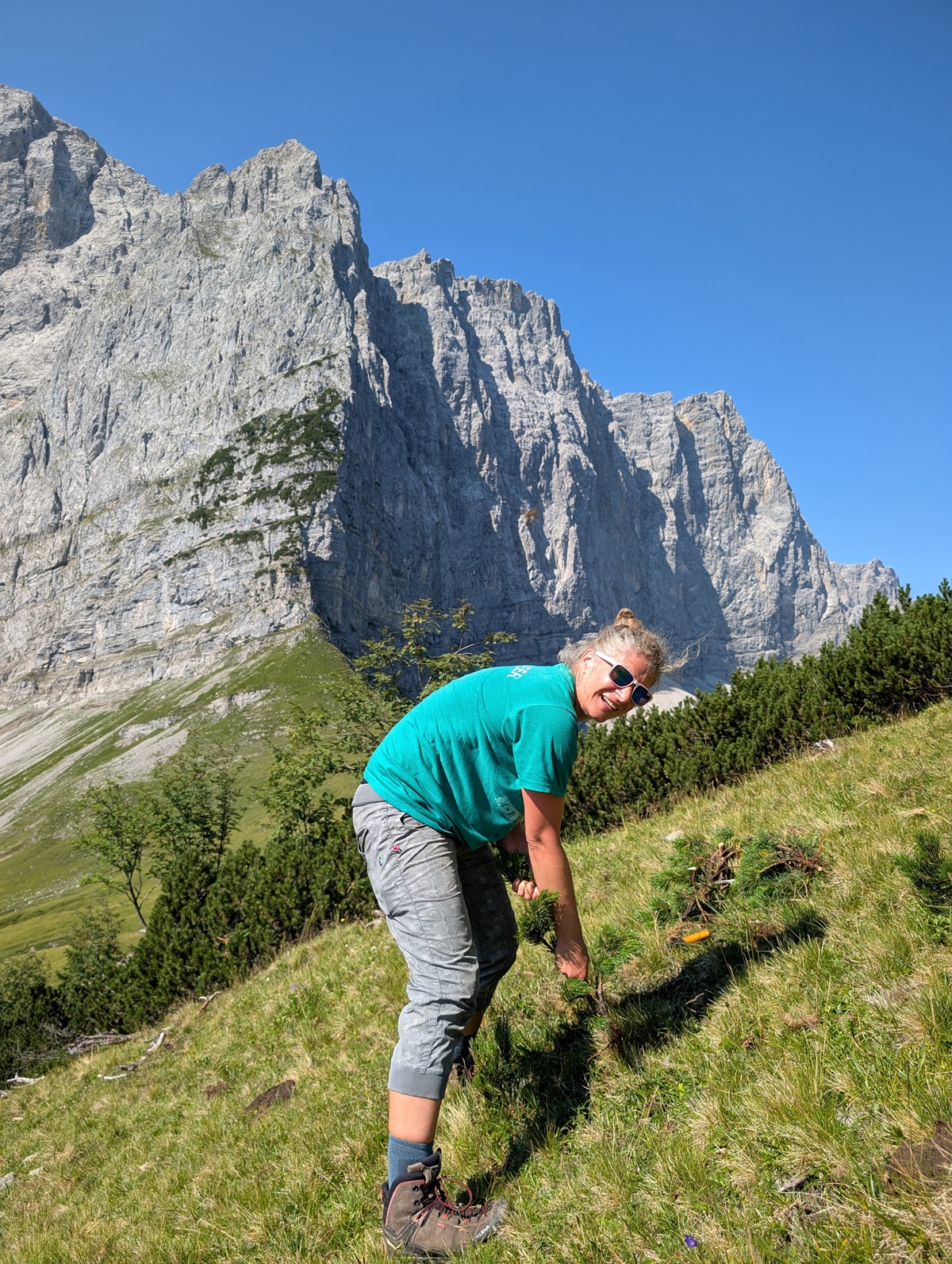 Eine Person mit Sonnenbrille und einem blaugrünen Hemd steht auf einem grasbewachsenen Hang, der zur Almpflege Lalidersalm gehört, mit felsigen Bergen im Hintergrund unter einem strahlend blauen Himmel.