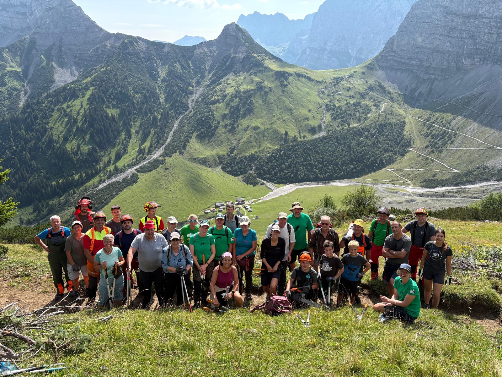 Eine Gruppe von Menschen posiert mit Wanderausrüstung auf einem grasbewachsenen Berghang in der Nähe der Lalidersalm, im Hintergrund sind unter einem teilweise bewölkten Himmel grüne Hügel, eine kurvenreiche Straße und hohe Gipfel zu sehen.
