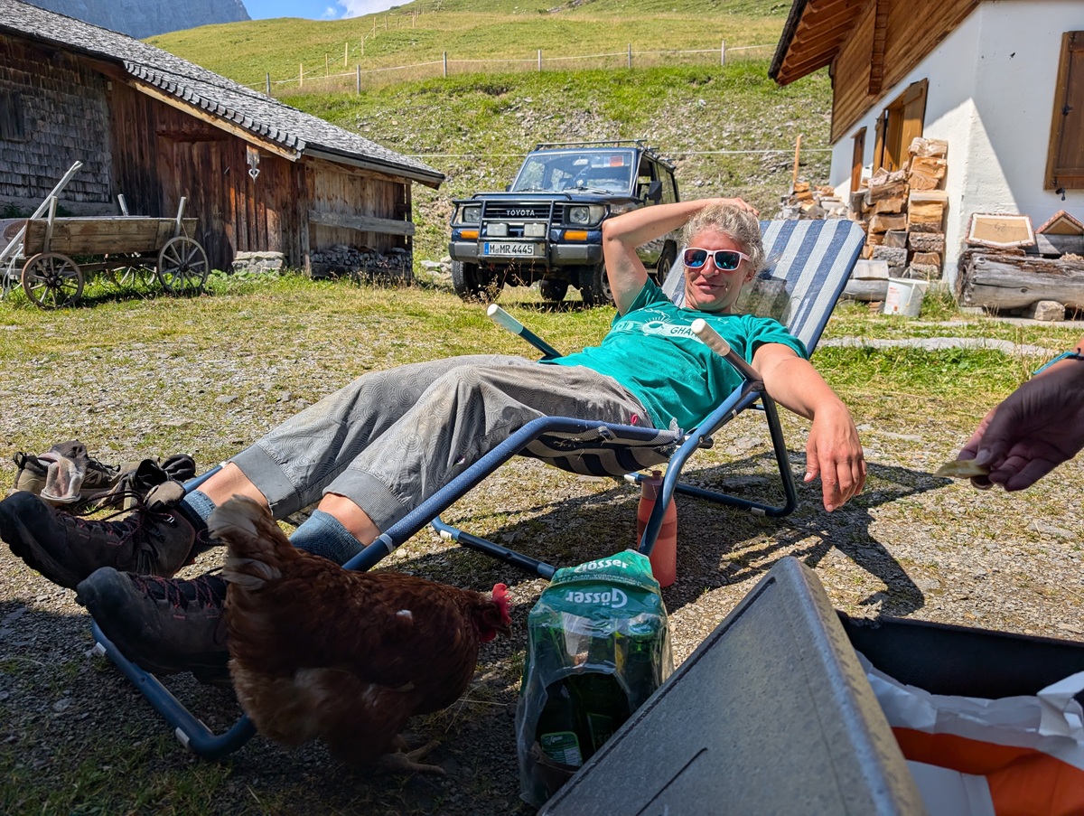 Eine Person, die sich im Freien auf einem Liegestuhl in der Nähe eines Holzhauses auf der Almpflege Lalidersalm entspannt, daneben ein Huhn und im Hintergrund ein geparktes Auto.