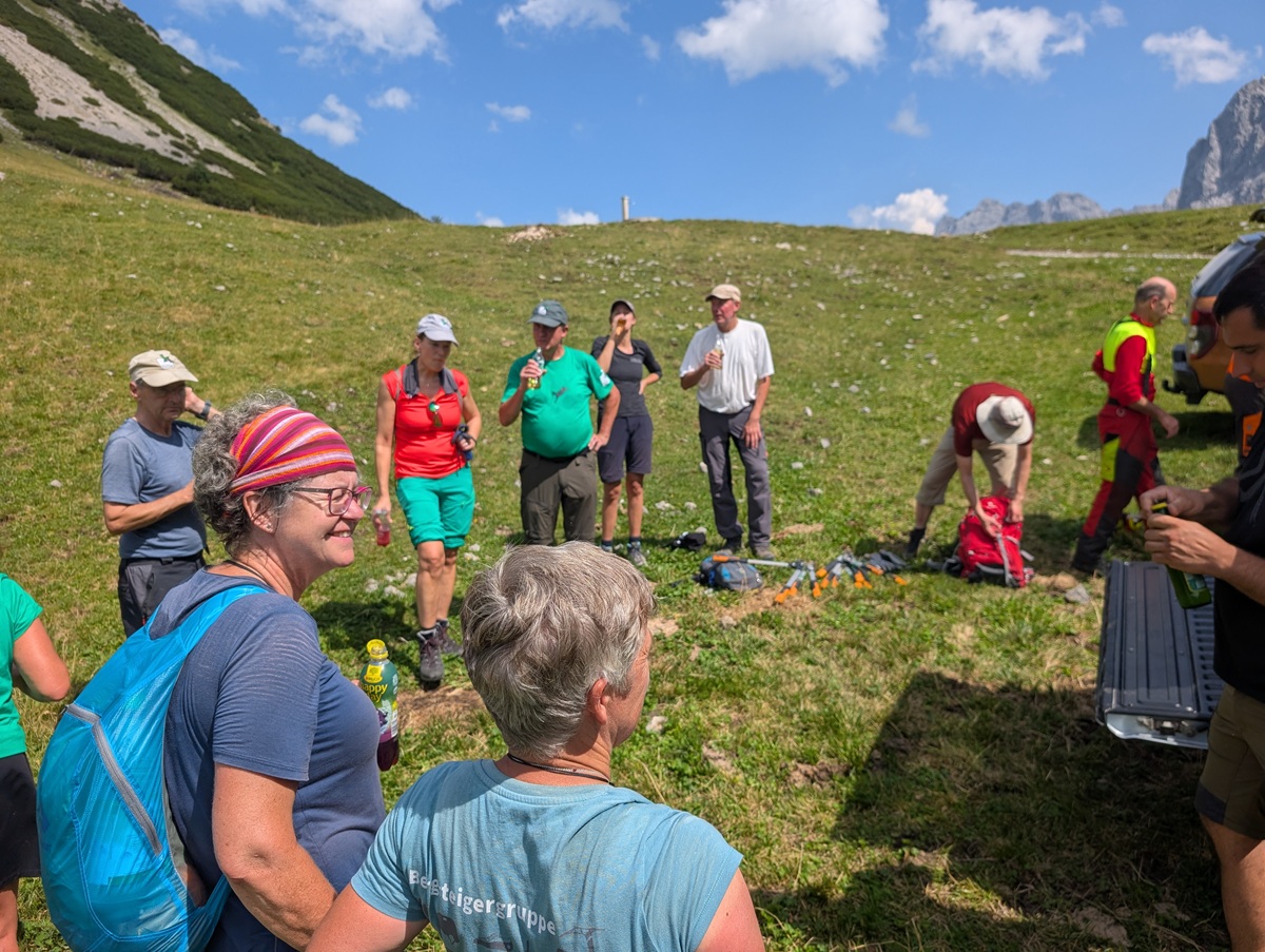 Auf der Lalidersalm steht und sitzt eine Gruppe von Wanderern unter blauem Himmel auf einem grasbewachsenen Hang und macht eine Pause, während ihre Getränke und Rucksäcke auf dem Boden verstreut liegen.