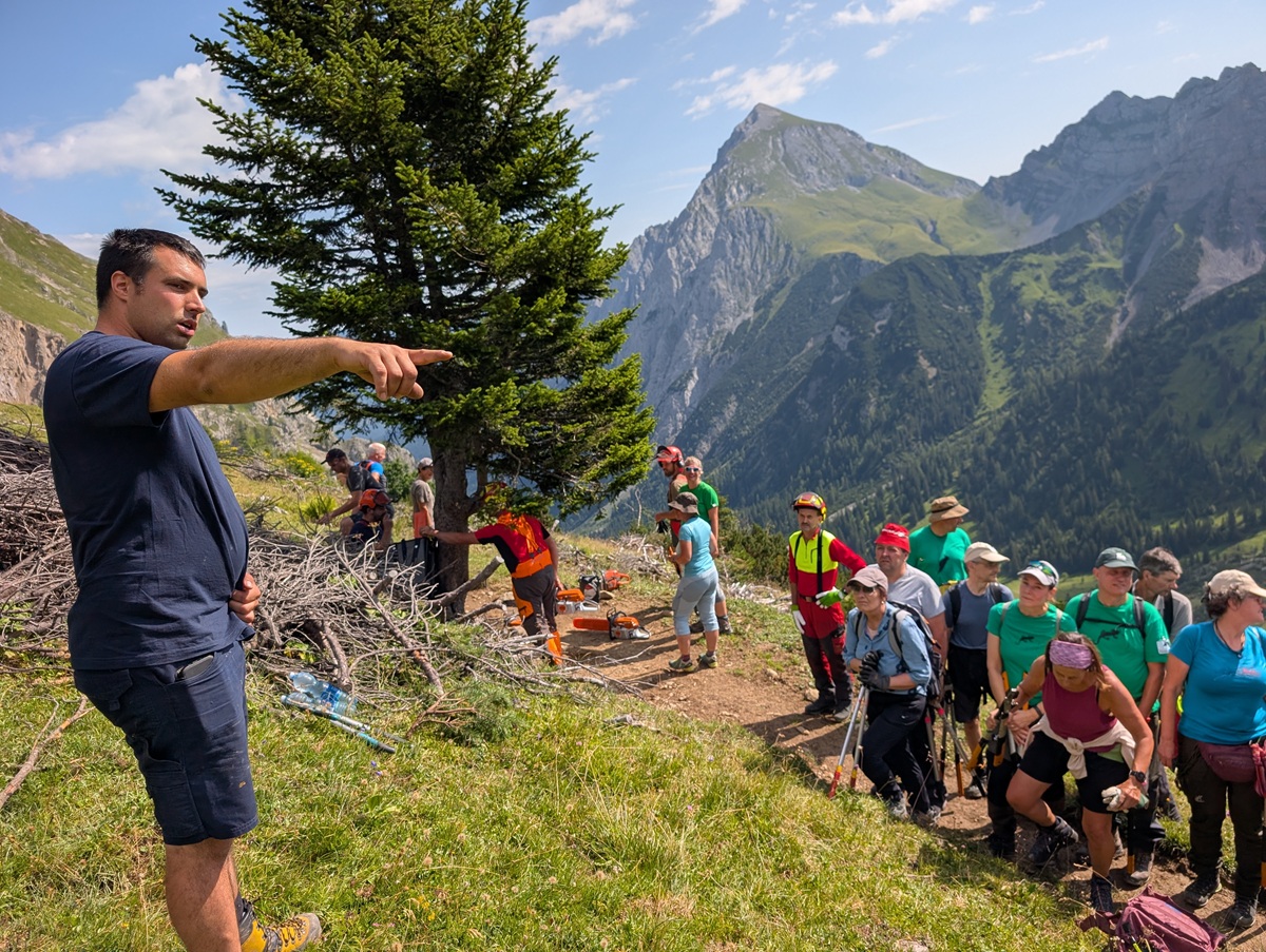 Ein Mann zeigt mit dem Finger in eine Richtung, während eine Gruppe von Wanderern mit Wanderstöcken auf einem Bergweg steht, umgeben von Bäumen und fernen Gipfeln unter einem teilweise bewölkten Himmel, und die malerische Route in der Nähe der Lalidersalm erkundet.