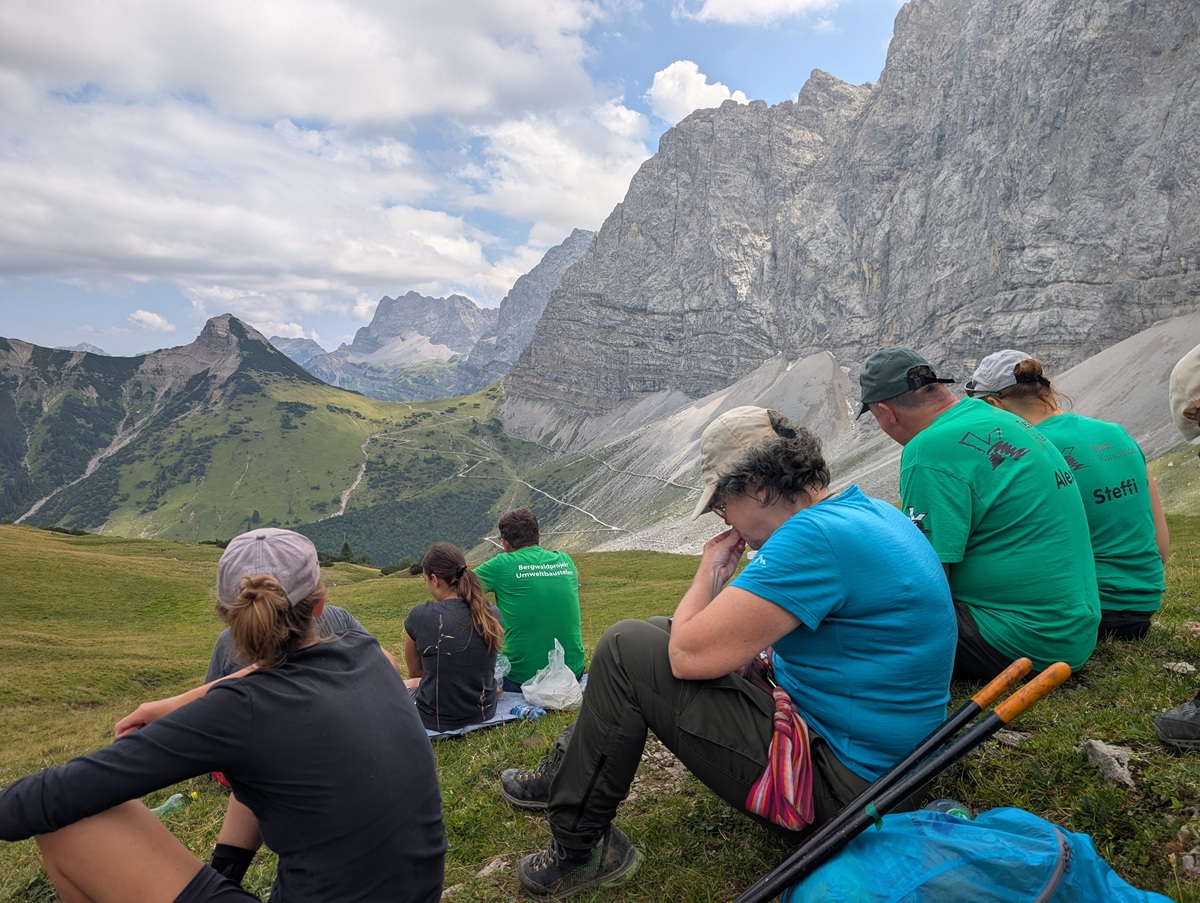 Eine Gruppe von Wanderern in legerer Outdoor-Kleidung sitzt auf einem grasbewachsenen Hang in der Nähe der Lalidersalm und ruht sich aus, im Hintergrund sind felsige Berge und ein bewölkter Himmel zu sehen.