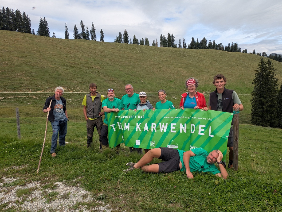Neun Personen stehen und eine liegt vor einem grünen „Team Karwendel“-Banner auf einem grasbewachsenen Hang, im Hintergrund sind Bäume und ein bewölkter Himmel zu sehen – ein Bild, das den Geist der Almpflege Lalidersalm und die Teamarbeit inmitten der Natur einfängt.