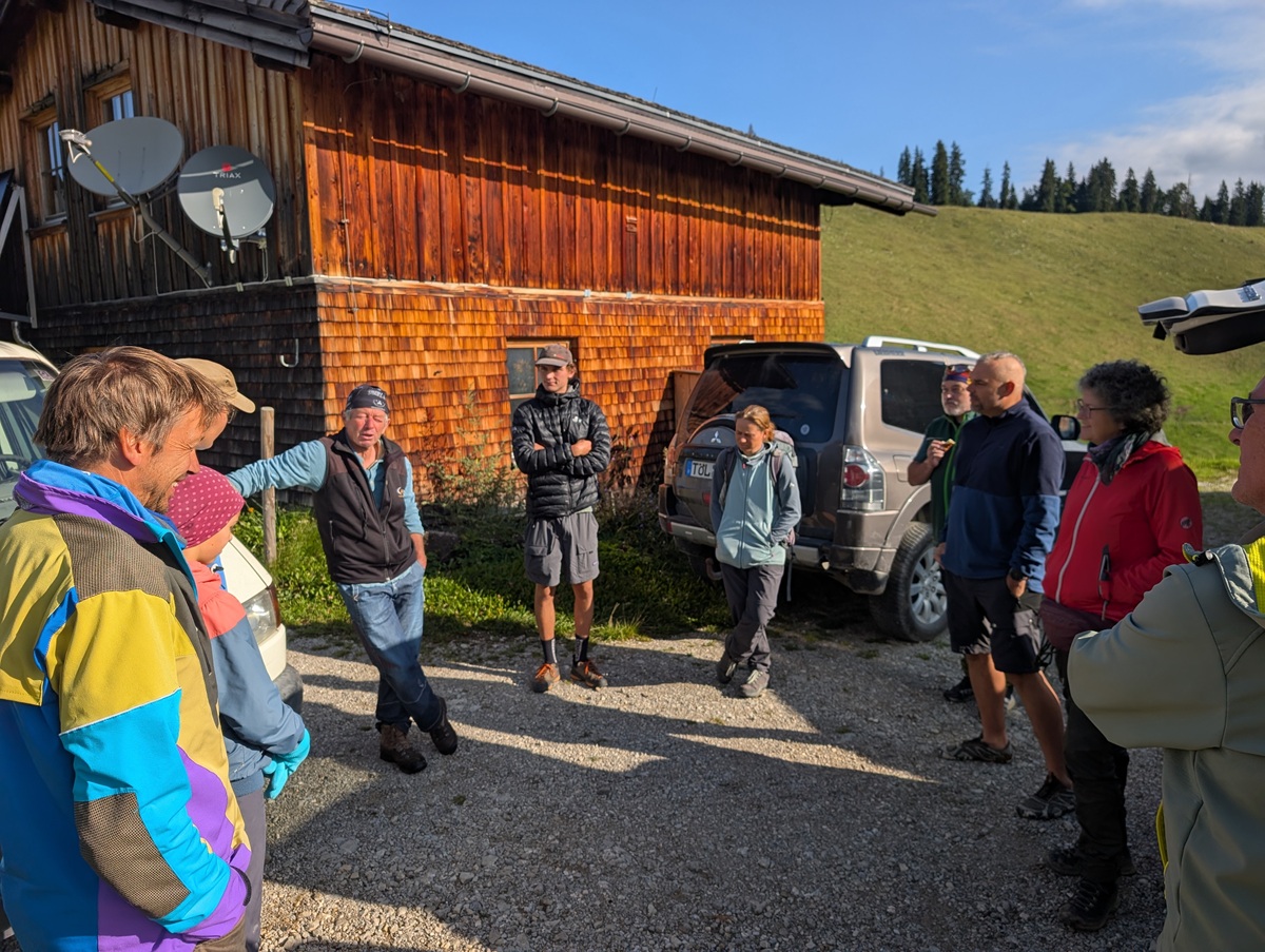 Eine Gruppe von Menschen steht im Freien in einem Kreis in der Nähe eines Holzgebäudes und geparkter Autos und unterhält sich im Sonnenlicht auf einer Schotterfläche während der Almpflege Lalidersalm.