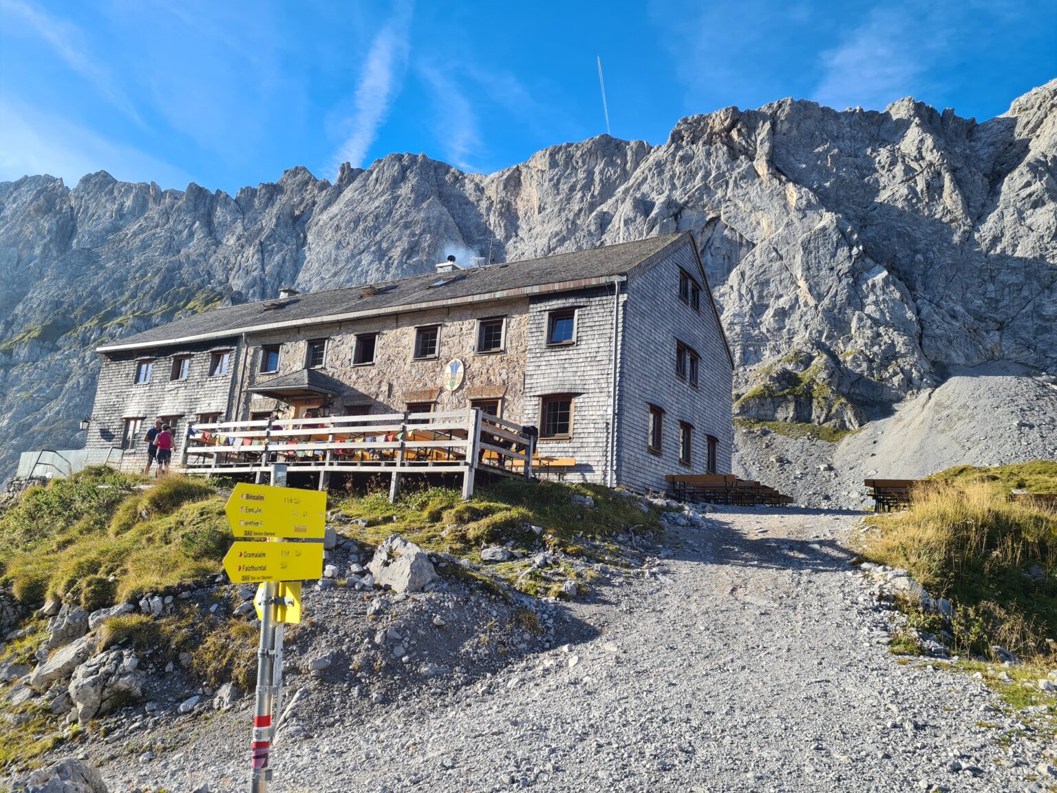 Am Fuße eines felsigen Gipfels in der Nähe der Walderalmteiche steht eine Berghütte mit einer Holzterrasse; im Vordergrund ist ein gelber Wanderwegweiser zu sehen.