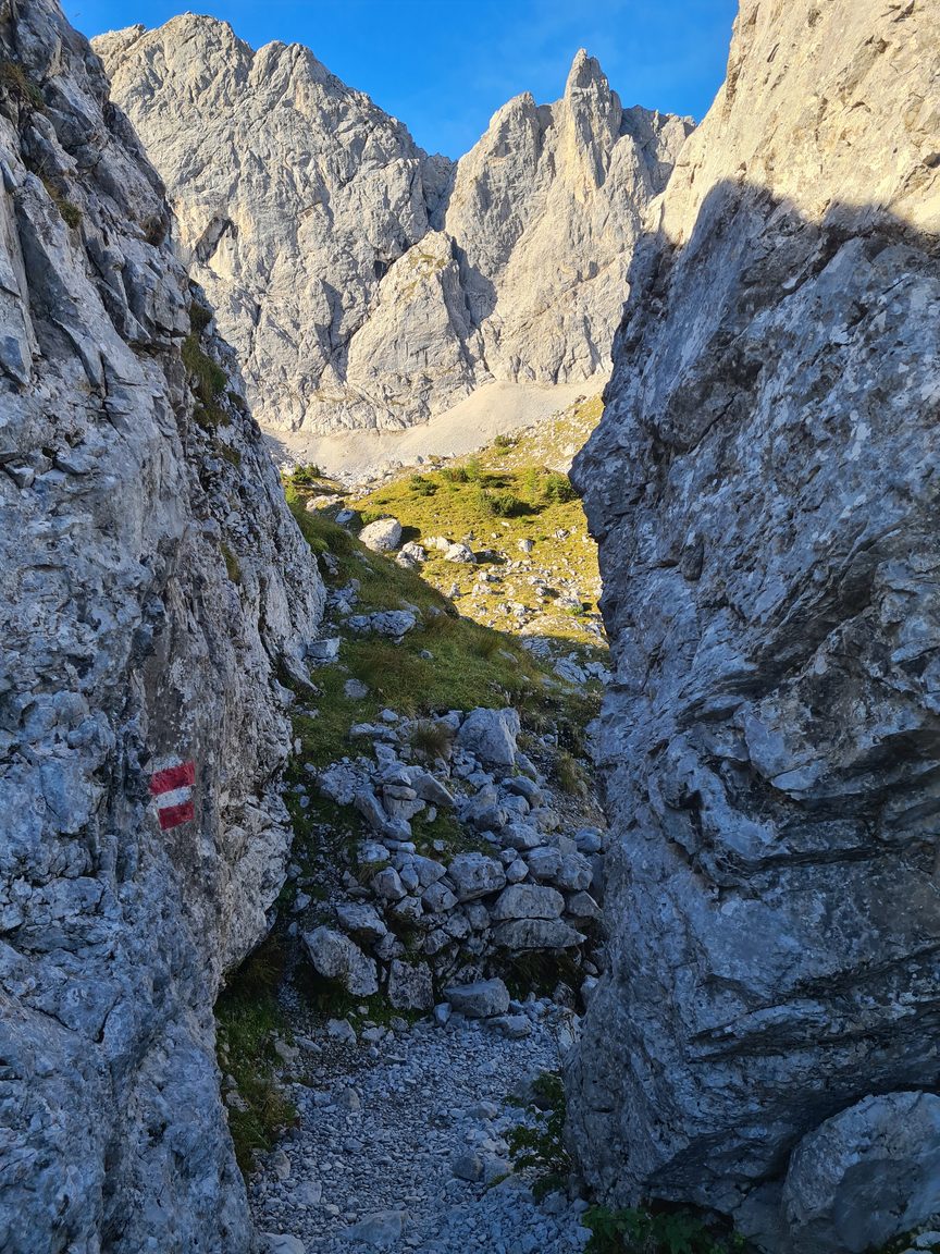 Ein felsiger Bergpfad in der Nähe der Walderalmteiche führt durch eine enge Lücke zwischen Felsblöcken, die durch ein rot-weißes Schild an der linken Felswand gekennzeichnet ist.