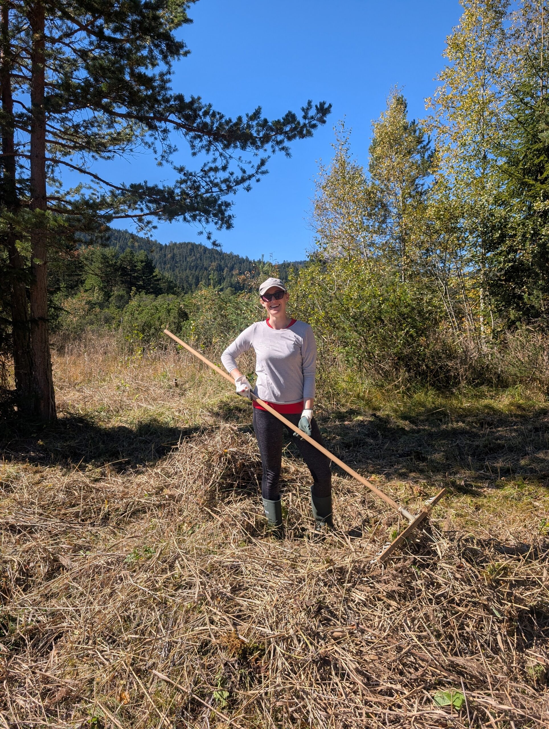 Eine Frau mit Hut und Sonnenbrille steht auf einer Wiese bei der Flachalm und hält einen Holzrechen in der Hand.