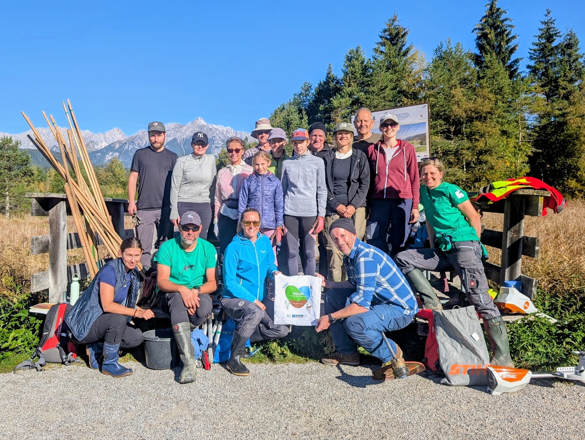 Eine große Gruppe von Freiwilligen posiert im Freien mit Ausrüstung und Werkzeugen und hält ein Schild für eine Wanderwegpflegeaktion von „Almpflege Flachalm“ in der Almenwelt Lofer hoch.