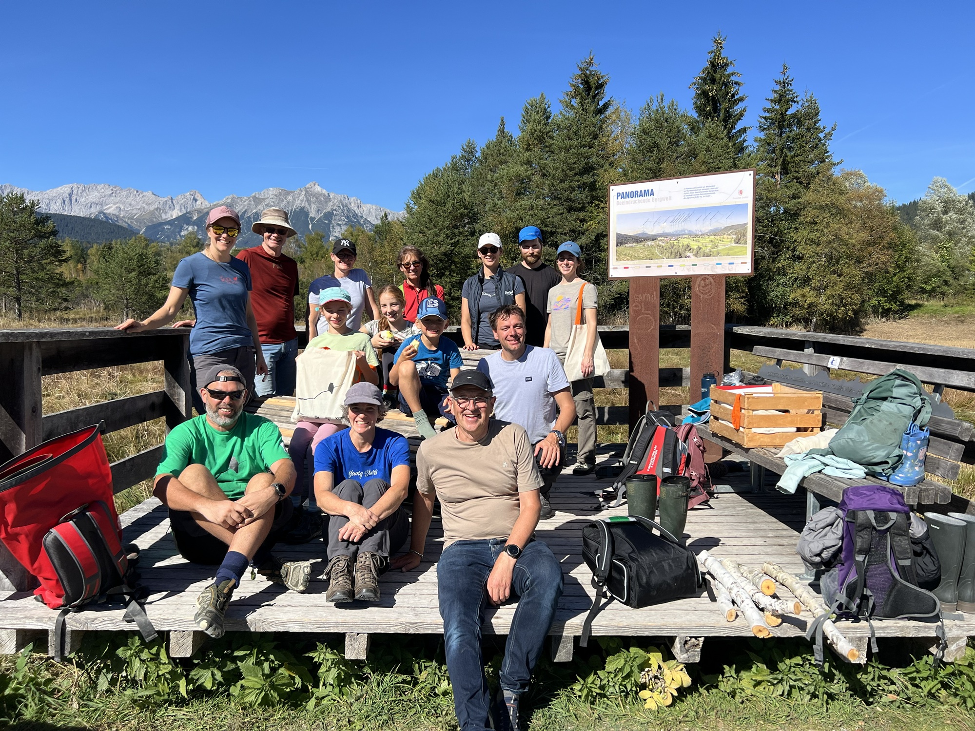 Eine Gruppe von Wanderern posiert für ein Foto auf einer hölzernen Aussichtsplattform bei der Almpflege Flachalm, im Hintergrund sind ein Panoramschild und Berge zu sehen.