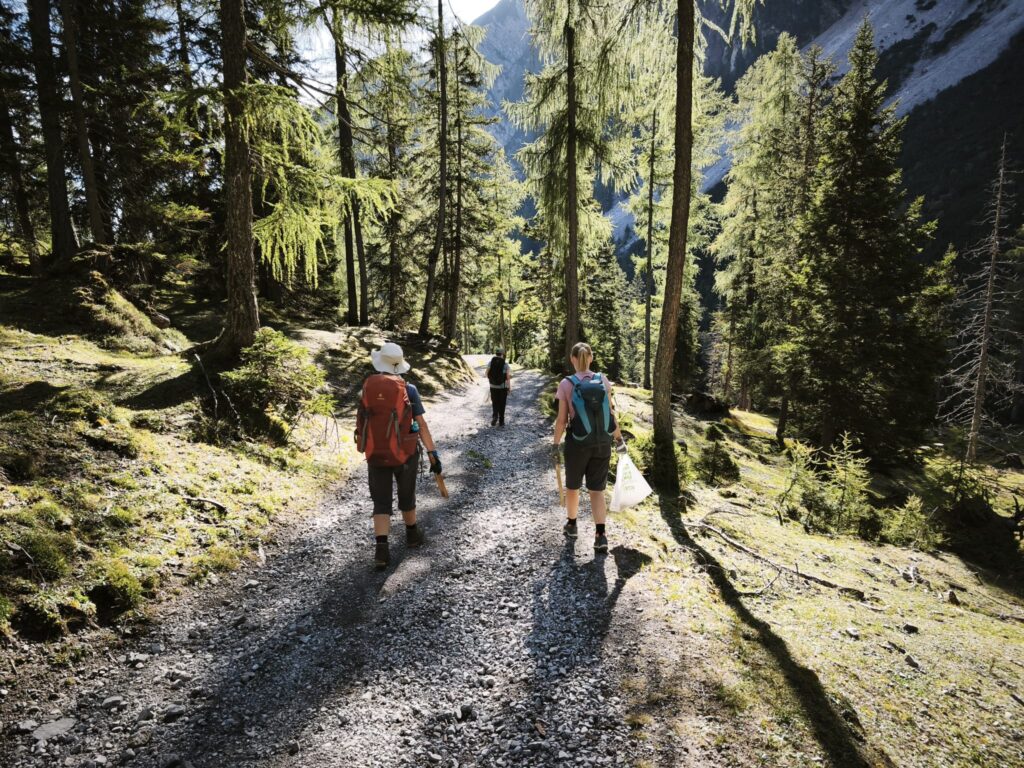 Drei Wanderer mit Rucksäcken gehen auf einem Waldweg mit Schotterbelag in der Nähe der Walderalmteiche entlang.
