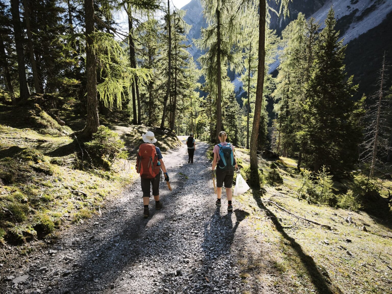 Drei Wanderer mit Rucksäcken gehen auf einem Waldweg mit Schotterbelag in der Nähe der Walderalmteiche entlang.