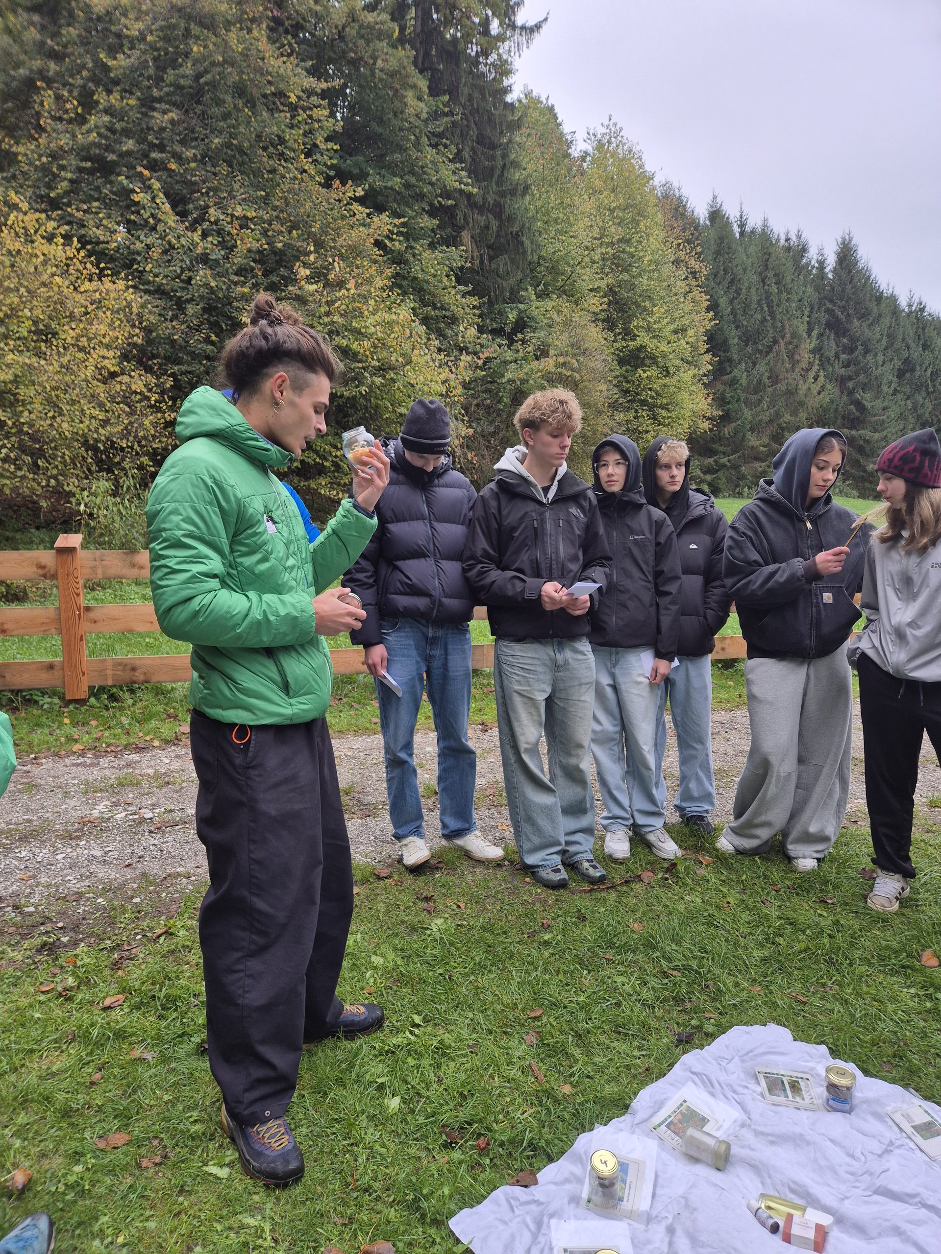 Ein junger Mann in einer grünen Jacke hält ein kleines Glas in der Hand, während er bei einer Müllsammelaktion auf einem grasbewachsenen Weg in der Nähe eines Waldes mit fünf Schülern spricht.